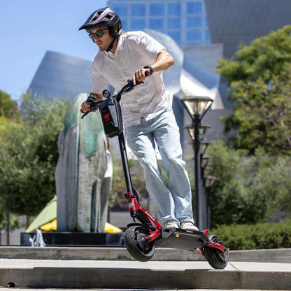 Person riding a red electric scooter in an urban setting with trees and buildings in the background.